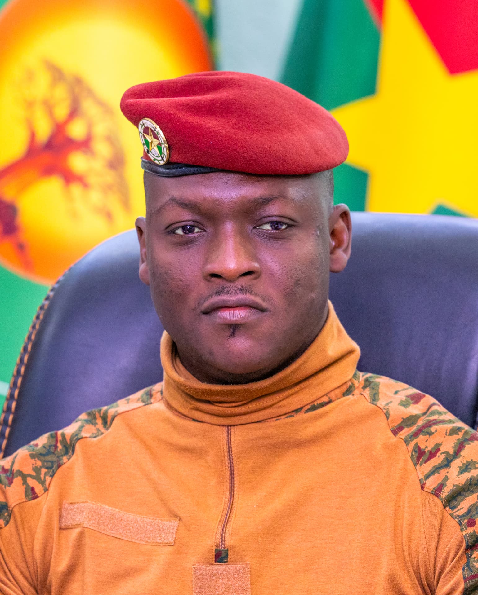 Portrait of Captain Ibrahim Traoré wearing a red beret, seated in front of the colours of the Burkina Faso flag.