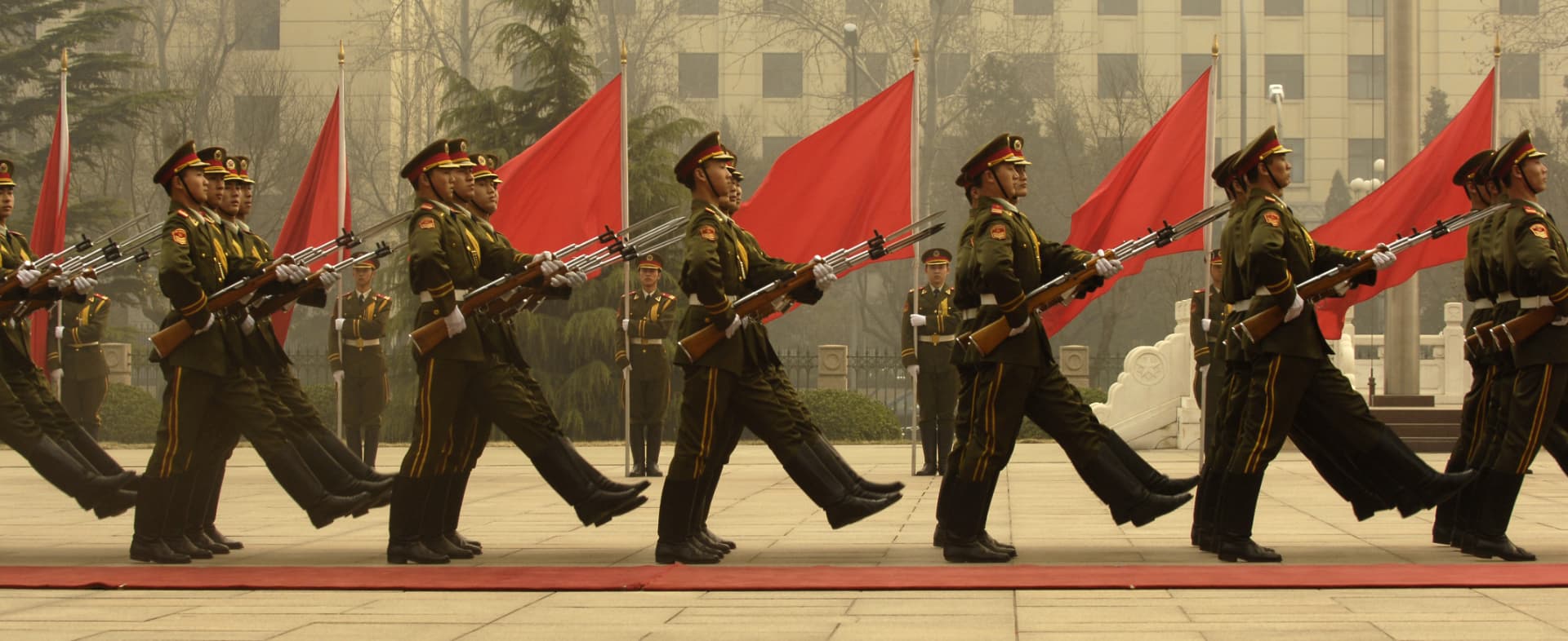 A line of soldiers marching in formation with rifles past red flags at a state ceremony in Beijing.